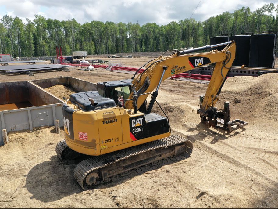 Yellow Caterpillar excavator on a construction site, digging into sand. Trees and blue sky in the background.