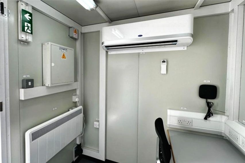 Interior of a small office. Air conditioner, heater, electrical panel, desk, and chair. Gray and white.