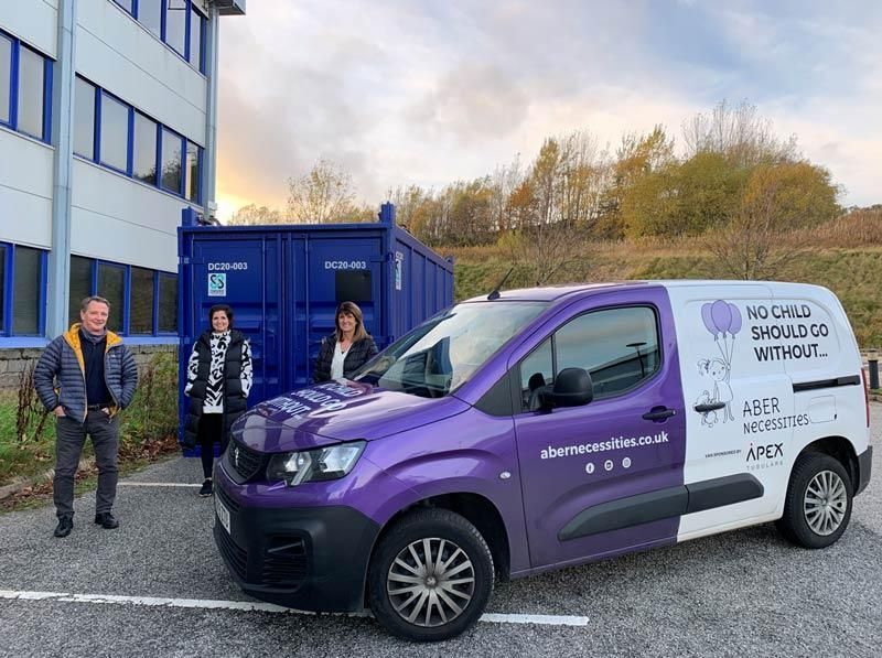 People standing beside a purple-and-white service van outside a building and blue containers