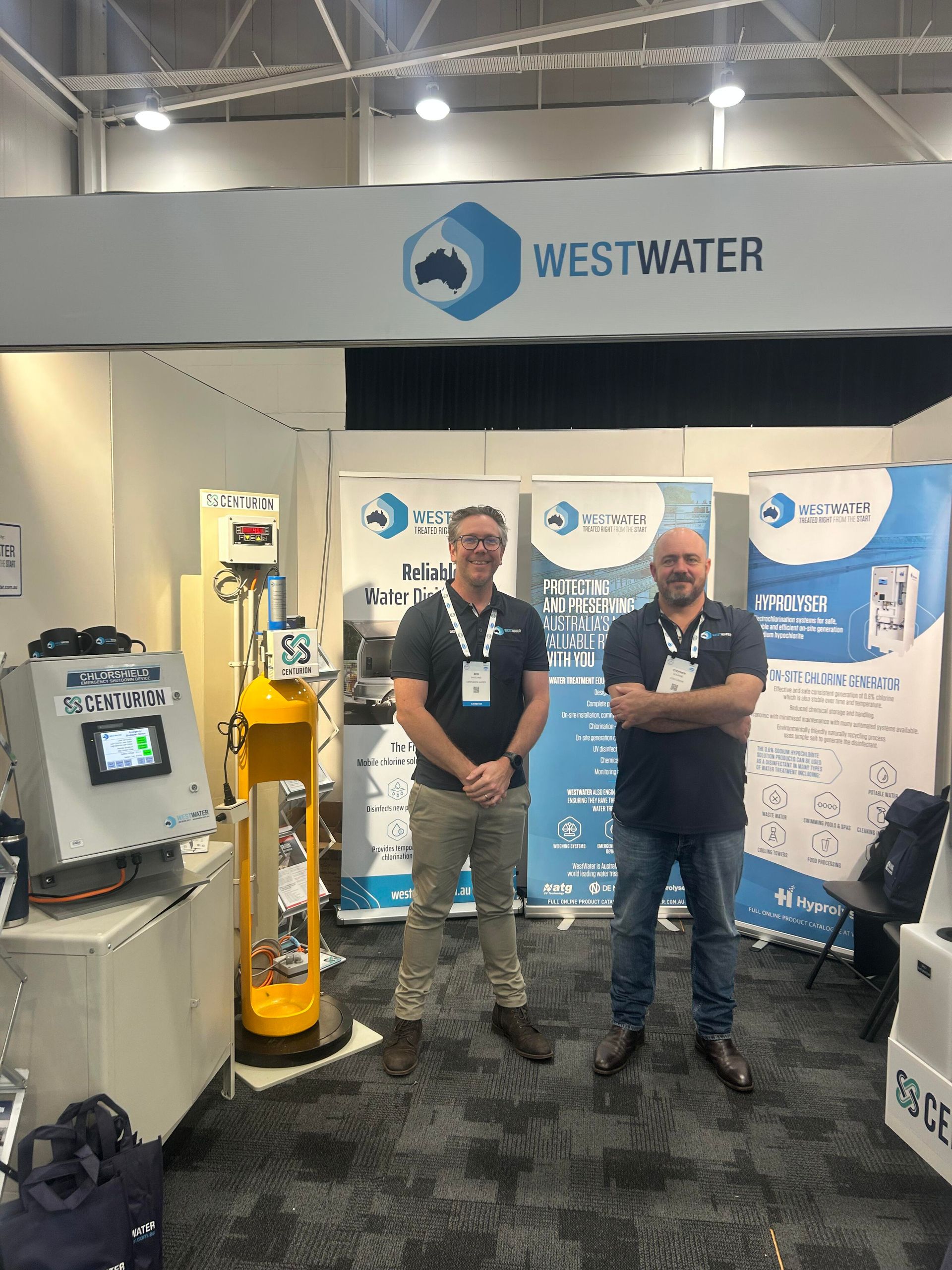 Two people standing at a Centurion WestWater booth at a trade show, with a large white and blue banner behind them and yellow and white chlorination equipment beside them.