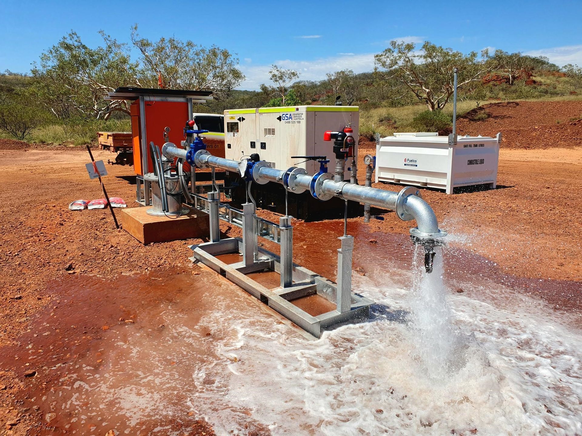 Pump station spraying water from metal pipes in a dry, red-soil landscape