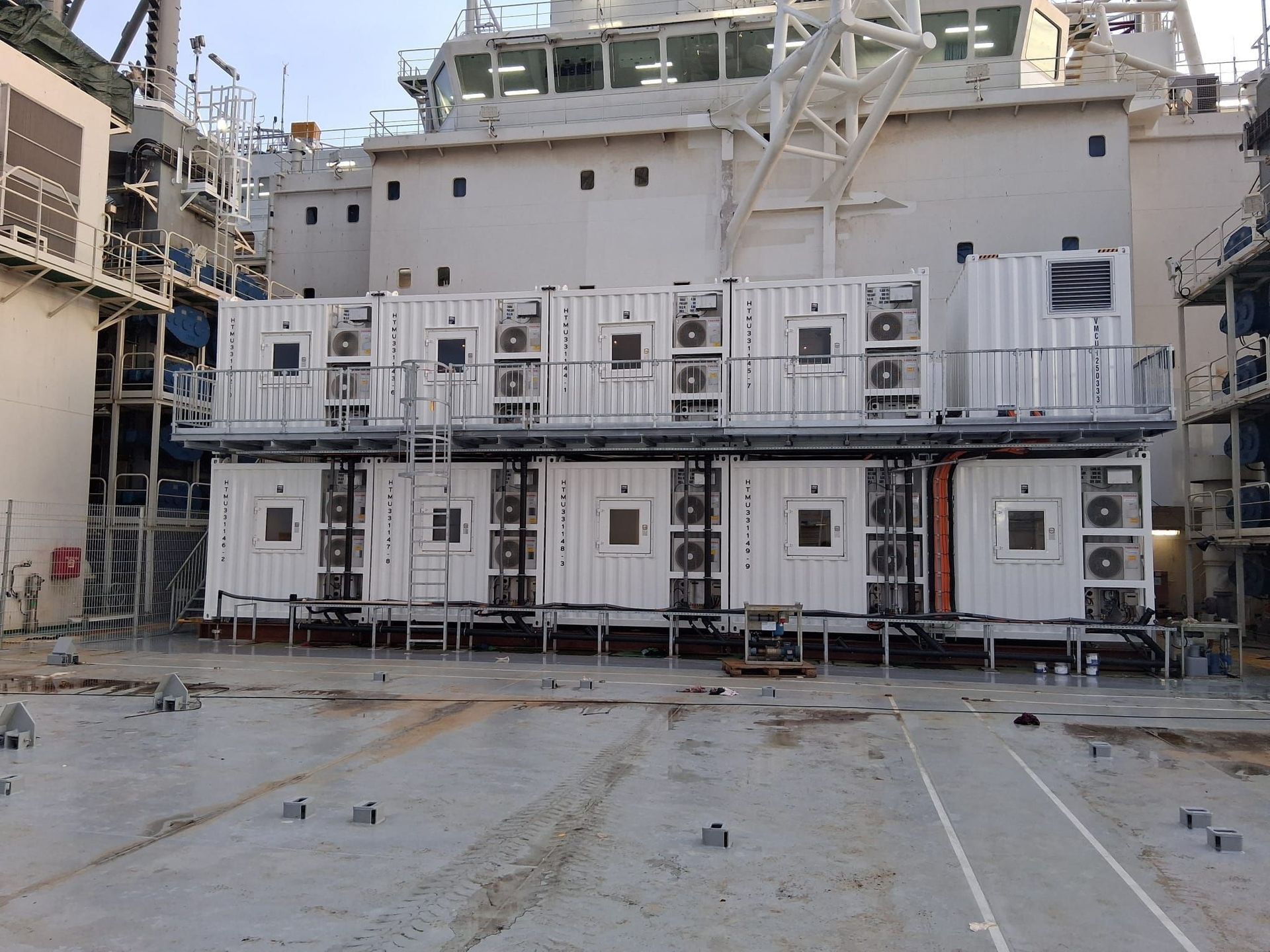 Industrial ship deck with stacked white container units and metal scaffolding on a wet concrete surface