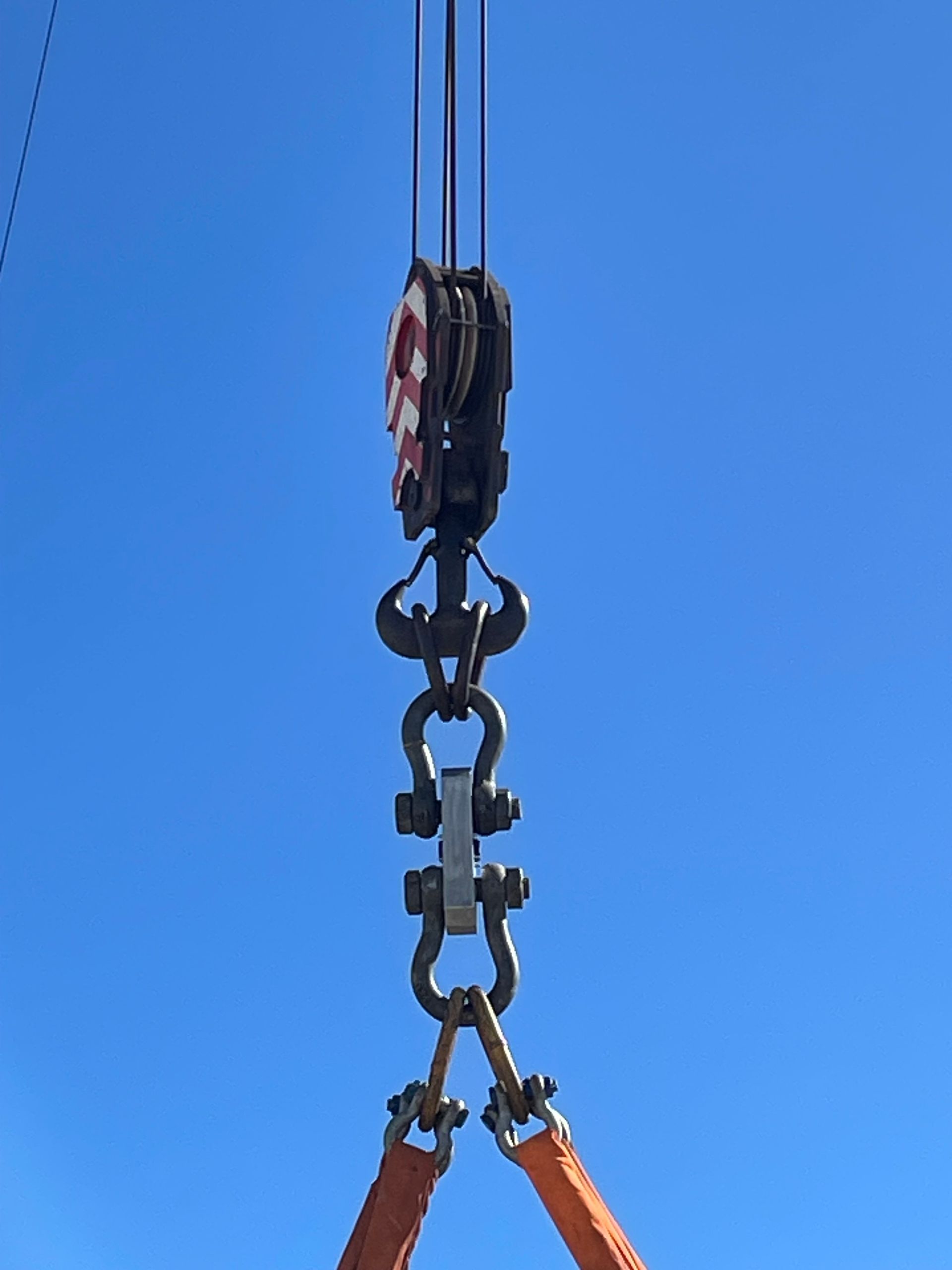 Crane hook with shackles and orange straps against a blue sky.