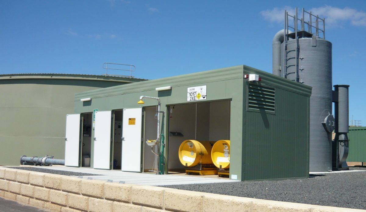 Green industrial building with open doors, two yellow tanks visible, and a large cylindrical structure behind it.
