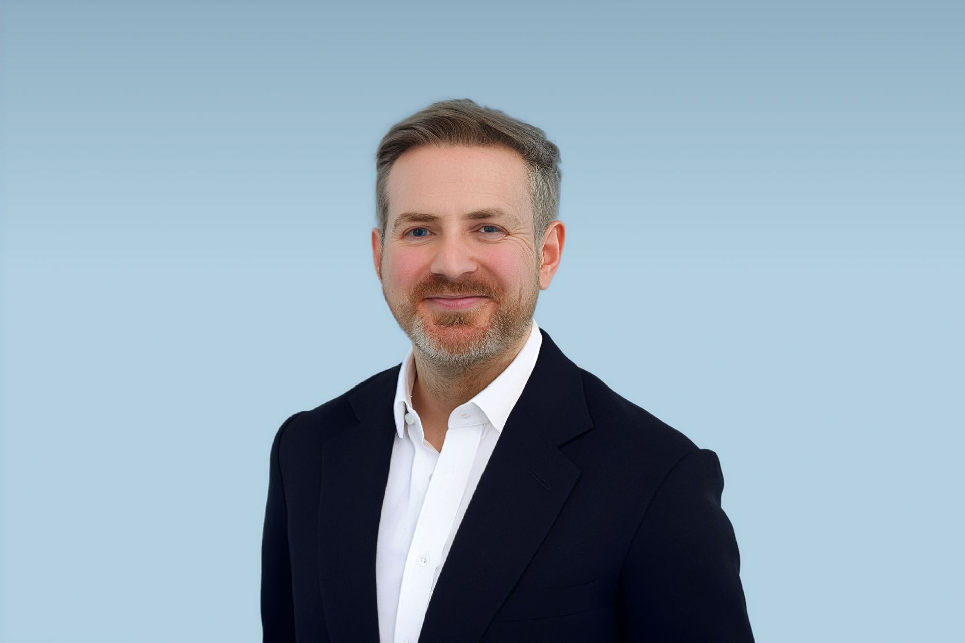 Man in a navy blazer and white shirt smiles, against a teal background.