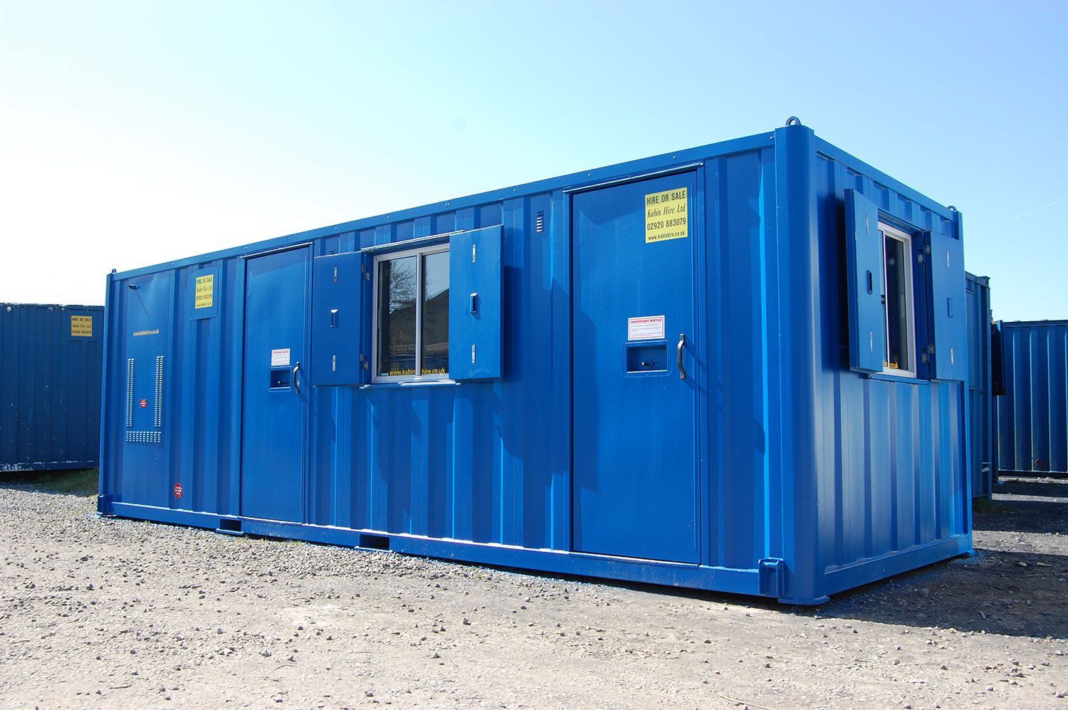 Blue metal container converted into an office, with windows and open shutters, outdoors on a sunny day.