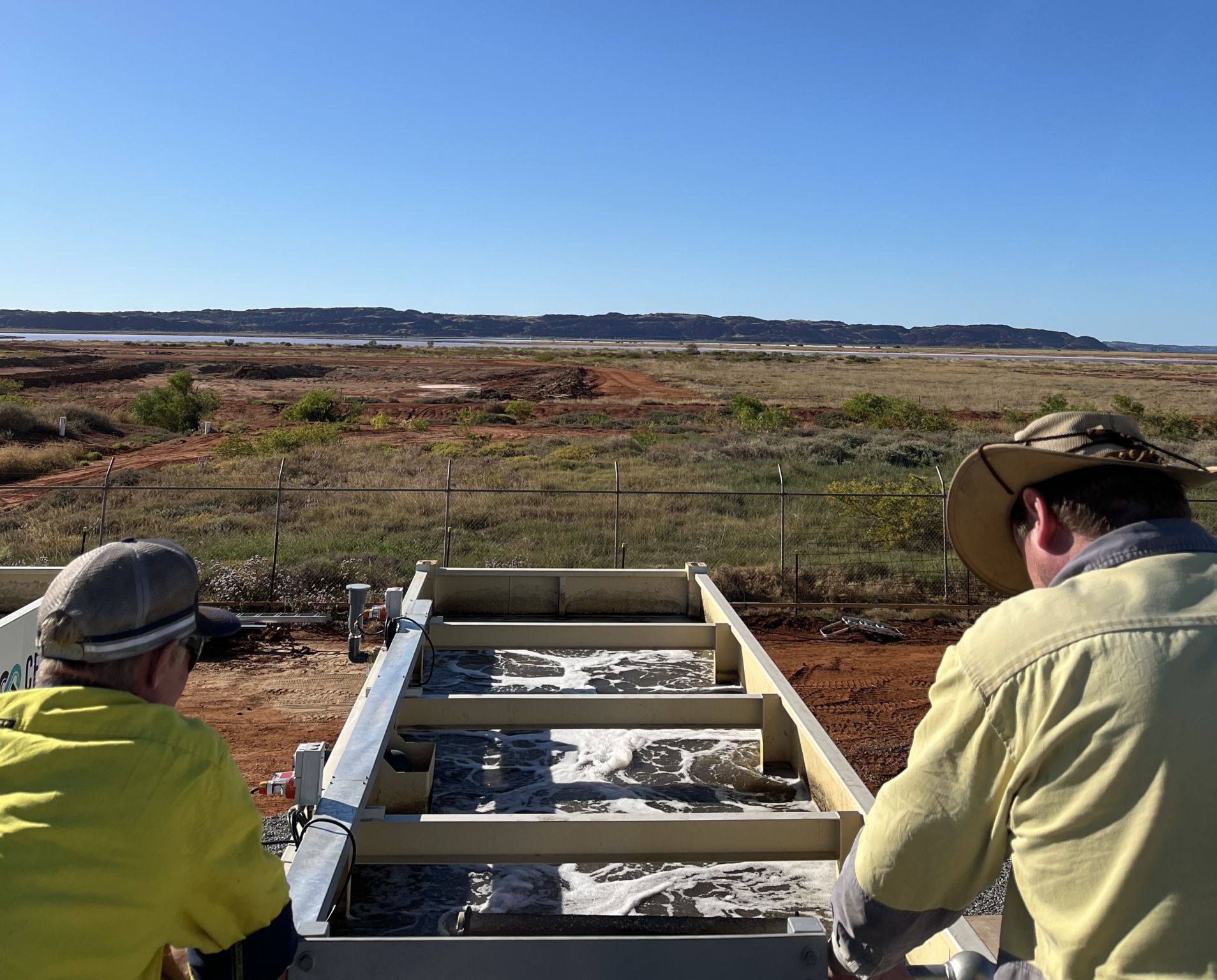 Two people in hats view a water treatment plant, open landscape under a clear blue sky.