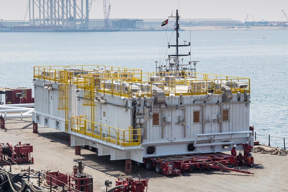 Large white and yellow industrial module on a trailer, near water and an offshore oil rig.