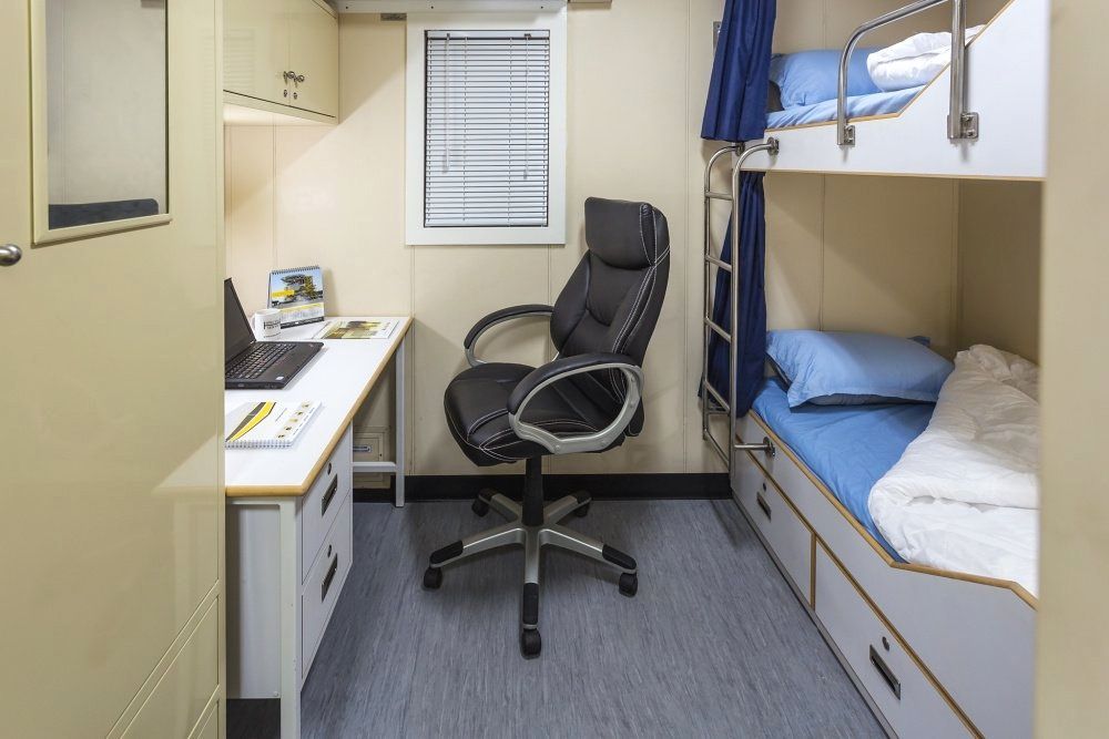 A small ship cabin with a desk, office chair, bunk beds, and a small window.