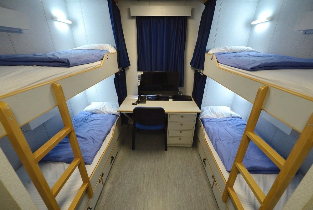 Four-bed bunkroom on a ship. Blue bedding and curtains, a desk with a computer. Lit by wall lamps.