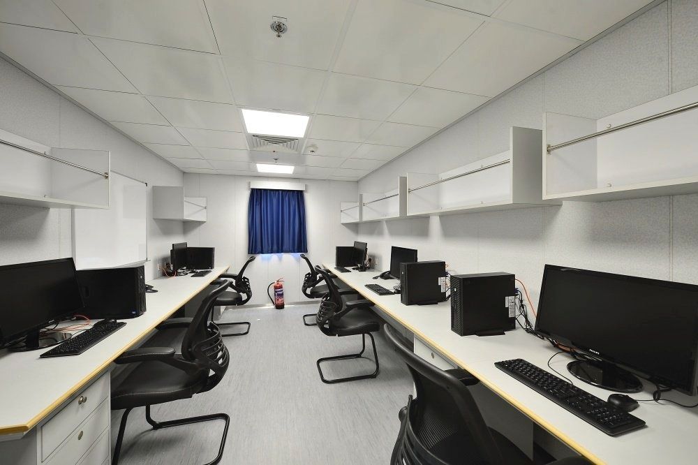 An office with computer desks, chairs, and monitors. White walls, cabinets, and ceiling. Blue curtain at the end.
