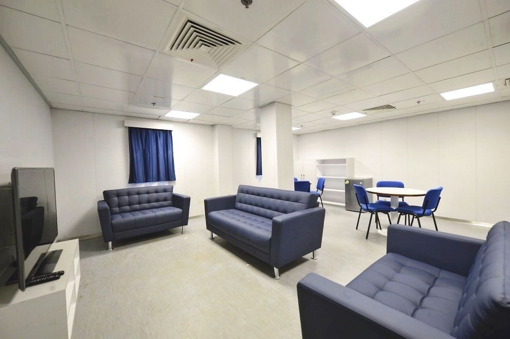 Lounge area with three blue sofas, a round table with blue chairs, and a TV. White walls and ceiling with blue curtains.