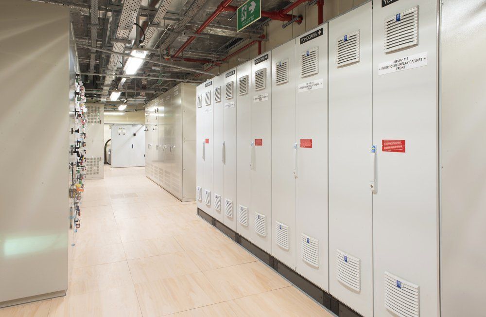 A narrow electrical room with rows of gray cabinets and overhead pipes. Light wood-colored floor.