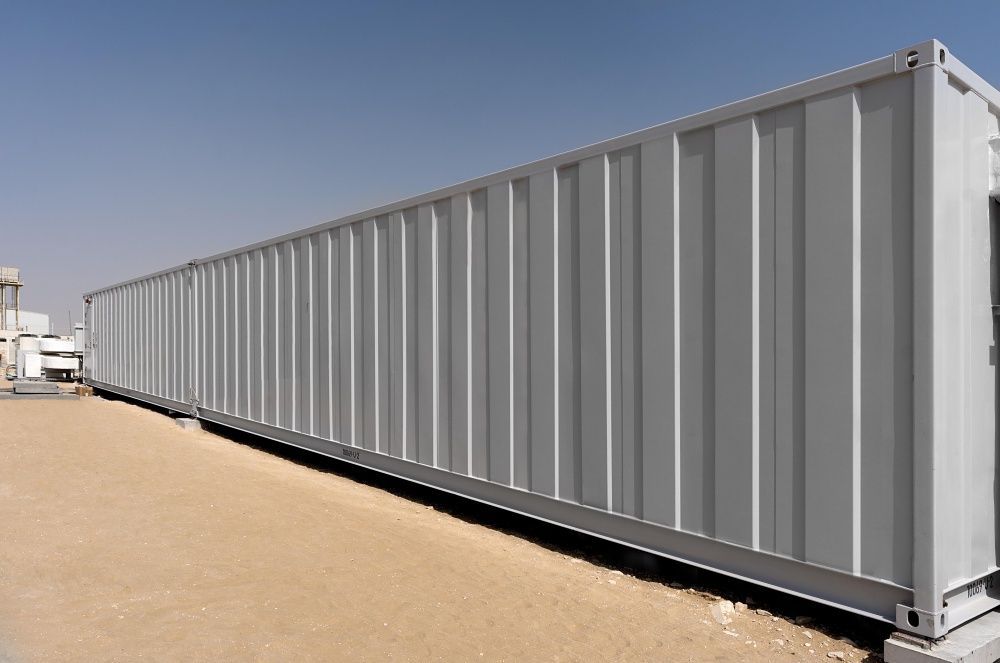 Long, light-gray shipping container on a tan surface against a clear blue sky.