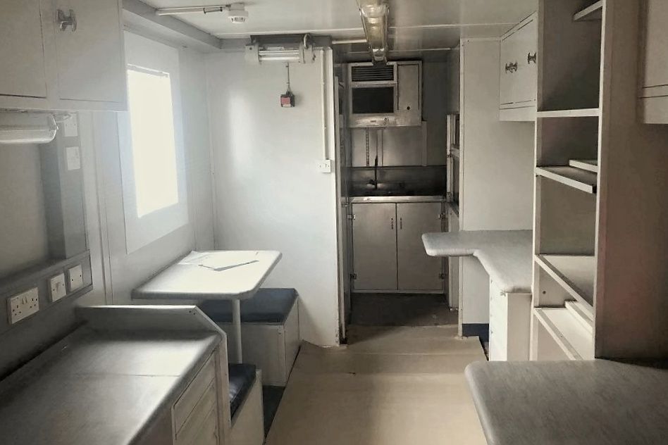 Interior of a small, narrow kitchen and dining area. White cabinets, stainless steel appliances, and a small table with bench seating.
