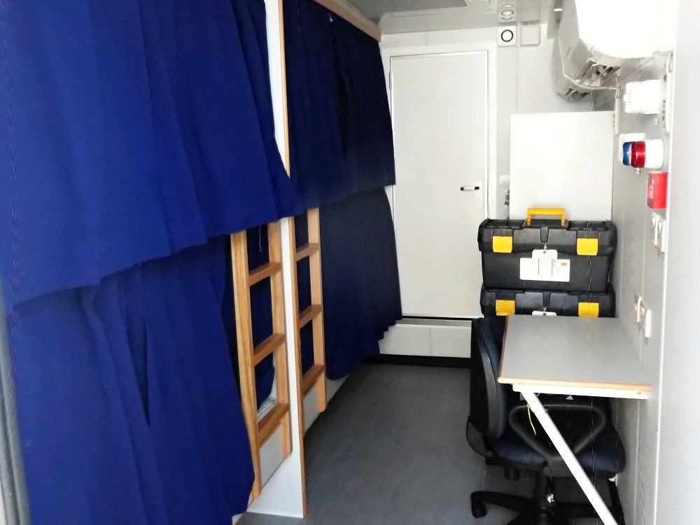 Interior of a mobile bunk room with two bunk beds and a desk, curtains, and a toolbox.