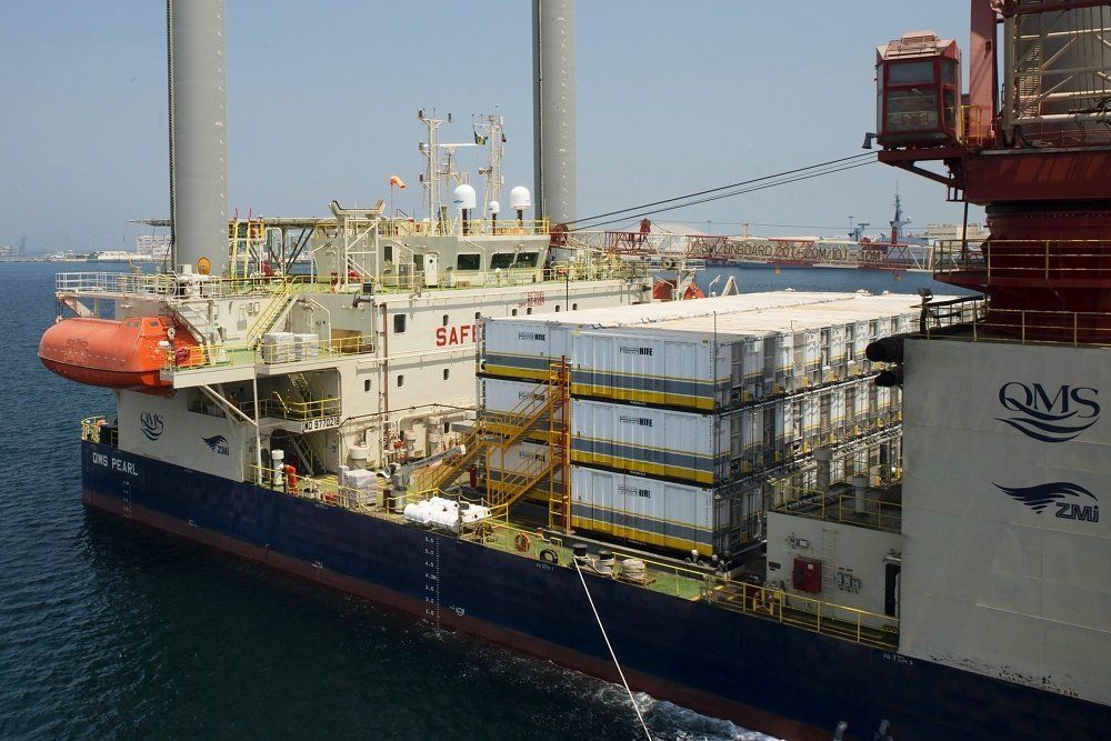 Blue and white ship with stacks of containers, docked near a red crane.