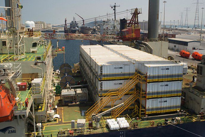 A cargo ship docked at a port with stacked white containers and a yellow staircase. Other ships and cranes are visible.