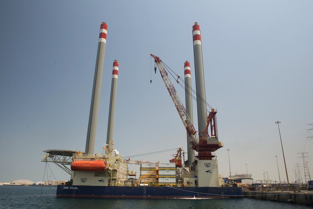 Offshore jack-up vessel in the water with four tall legs, a large crane, and containers, against a blue sky.