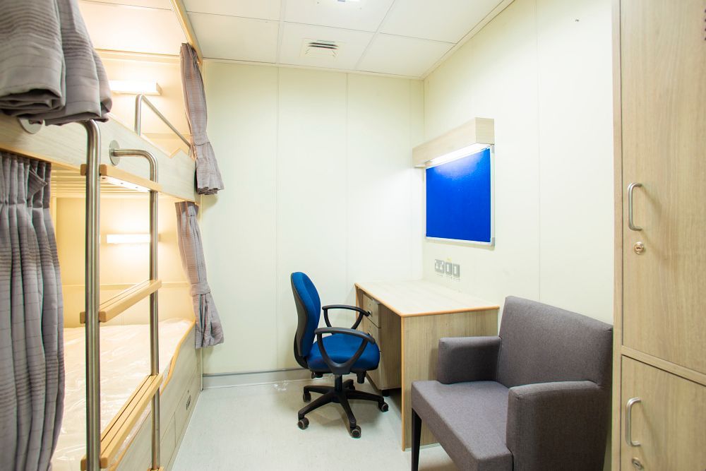 A small, beige room with bunk beds, desk, blue chair, and grey armchair.
