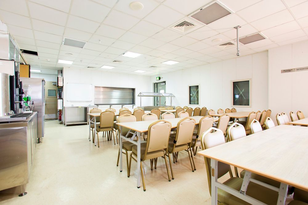 Empty cafeteria with tables, chairs, stainless steel kitchen equipment, and bright overhead lighting.