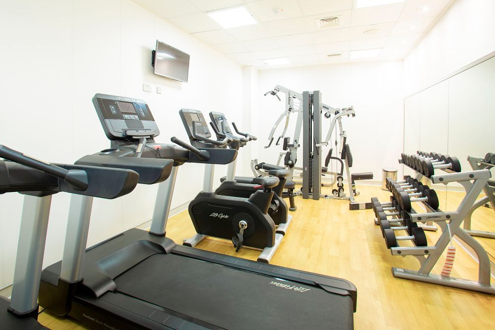 Gym with treadmills, stationary bike, weight machines, and dumbbells. Bright room, wooden floor, and a TV.
