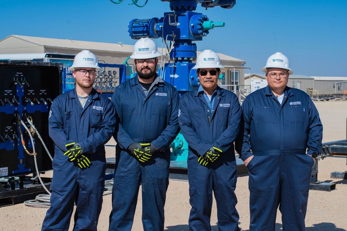 Four oil workers in blue jumpsuits and hard hats stand in front of oil well equipment.
