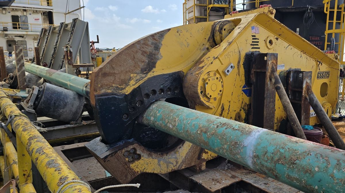 A large yellow cutting tool grips a green pipe on an offshore platform.