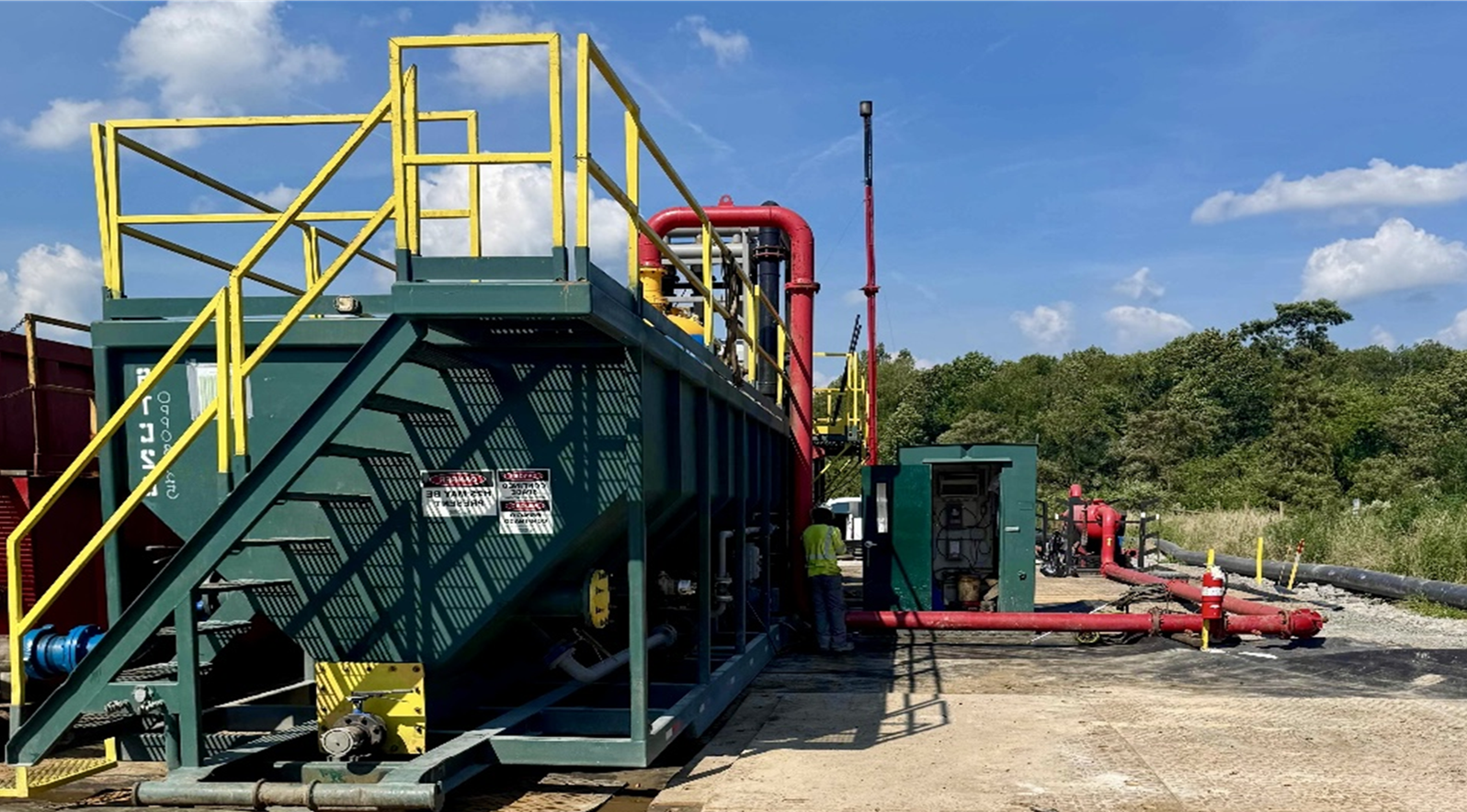 Green industrial equipment with yellow stairs and red piping on a gravel lot under a blue sky.