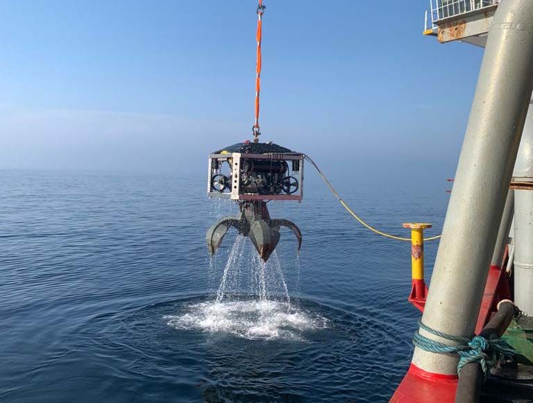 An underwater robot being lowered into the ocean from a ship; water sprays from the robot's claws.