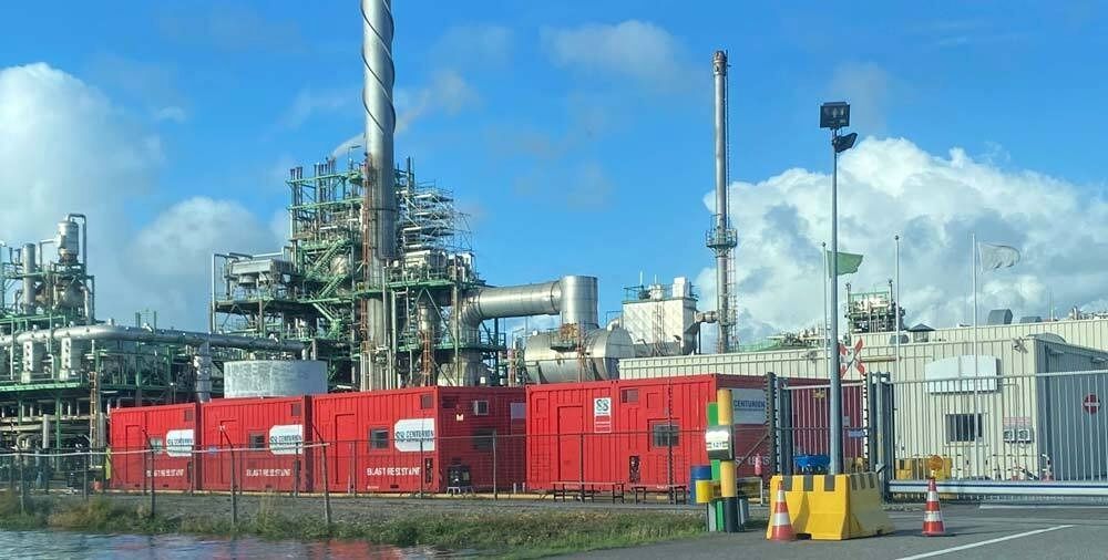 Industrial refinery with red containers, multiple pipes, and tall smokestacks against a blue sky.