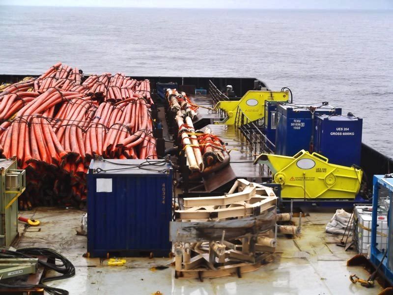 Deck of a ship with orange tubes, blue containers, yellow equipment, and the ocean.