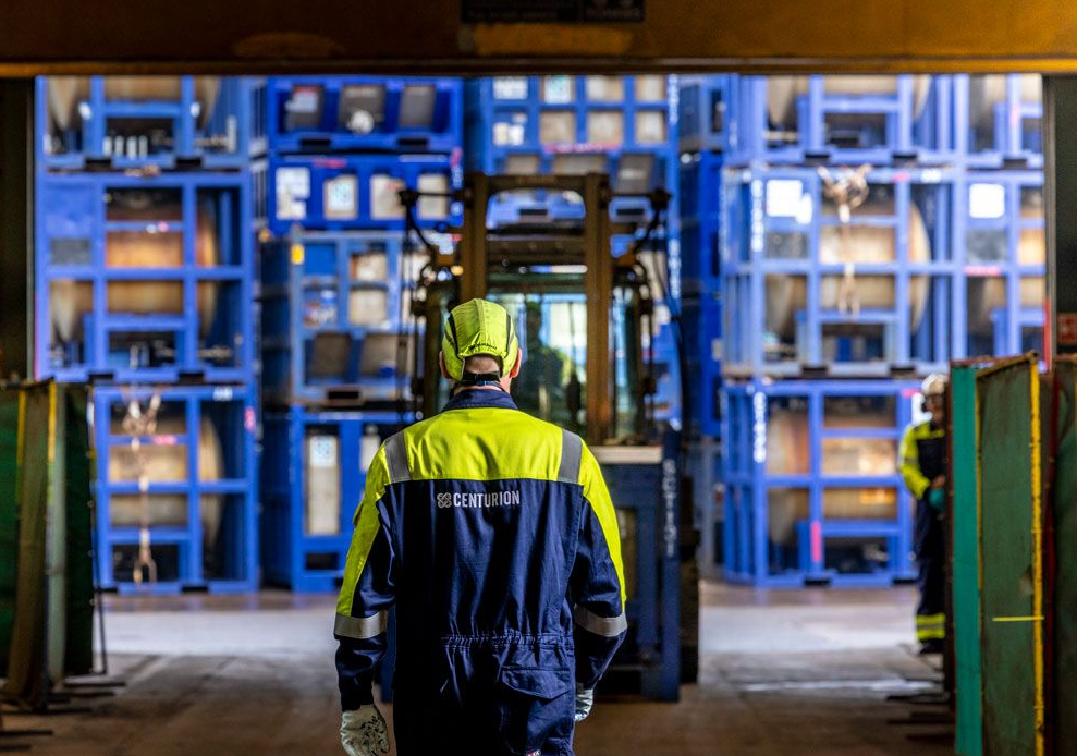 Worker in blue and yellow workwear walks toward a forklift in a warehouse with blue storage containers.