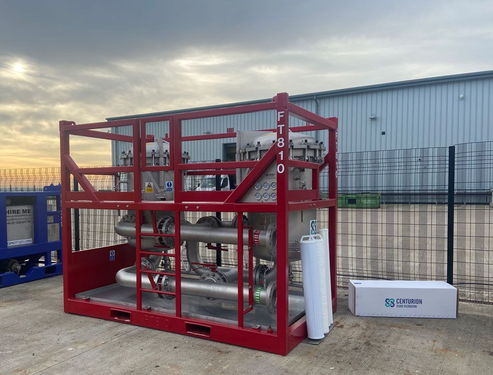 Red metal equipment cage with pipes and tanks, on a concrete surface outside with a blue container and white box.
