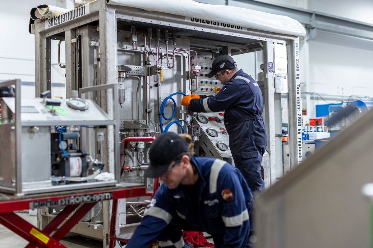 Two workers in blue coveralls and caps, working on industrial equipment.