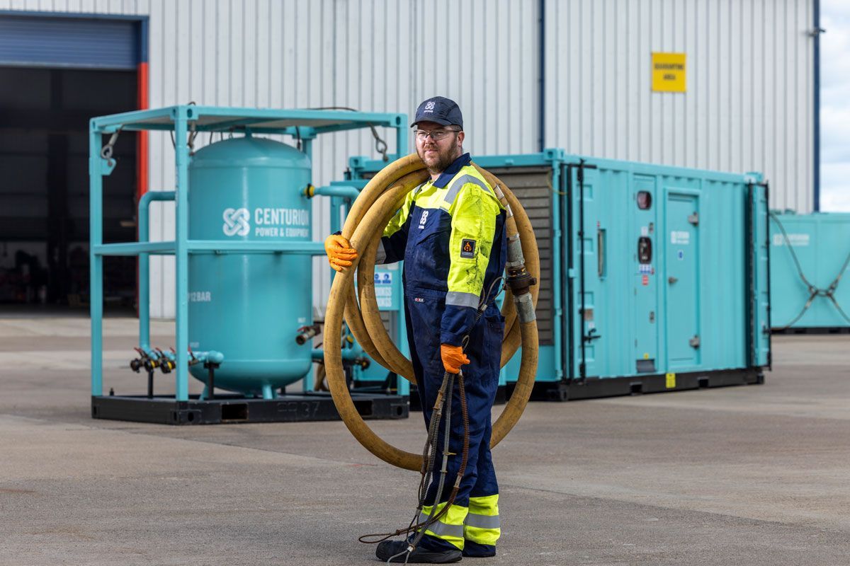 Worker holding hose next to industrial equipment in an outdoor setting.