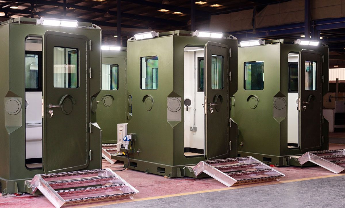 Three olive-green guard booths with open doors, each with a ramp. Industrial setting with overhead lights.