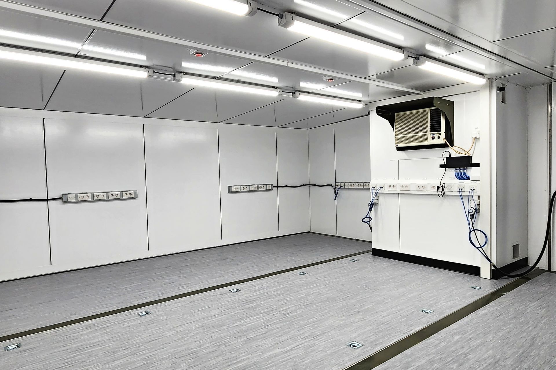 Interior of a white room with gray flooring. Fluorescent lights on the ceiling, a white cabinet, and electrical outlets along the walls.