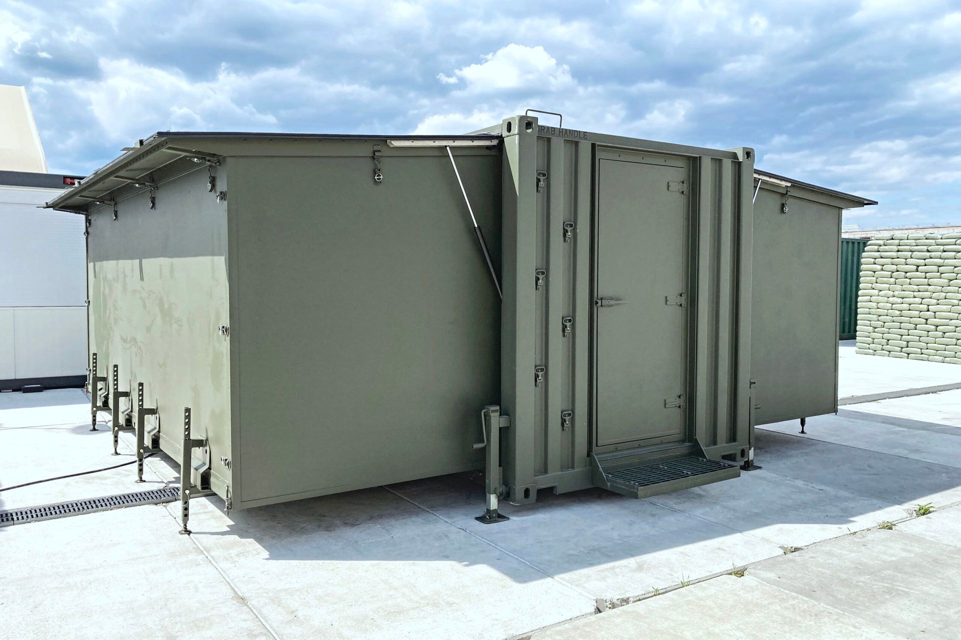 Olive green shelter with door and steps on a concrete surface, cloudy sky in background.