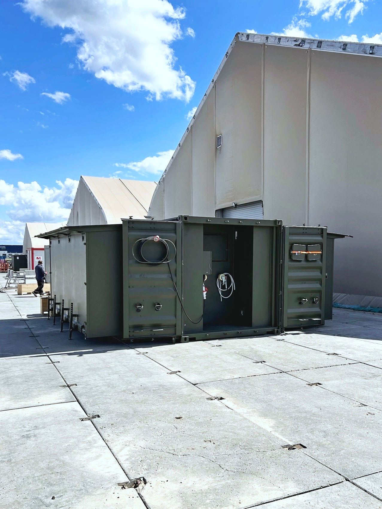 A green, modified shipping container with open doors sits outside a large building on a concrete surface under a blue sky.