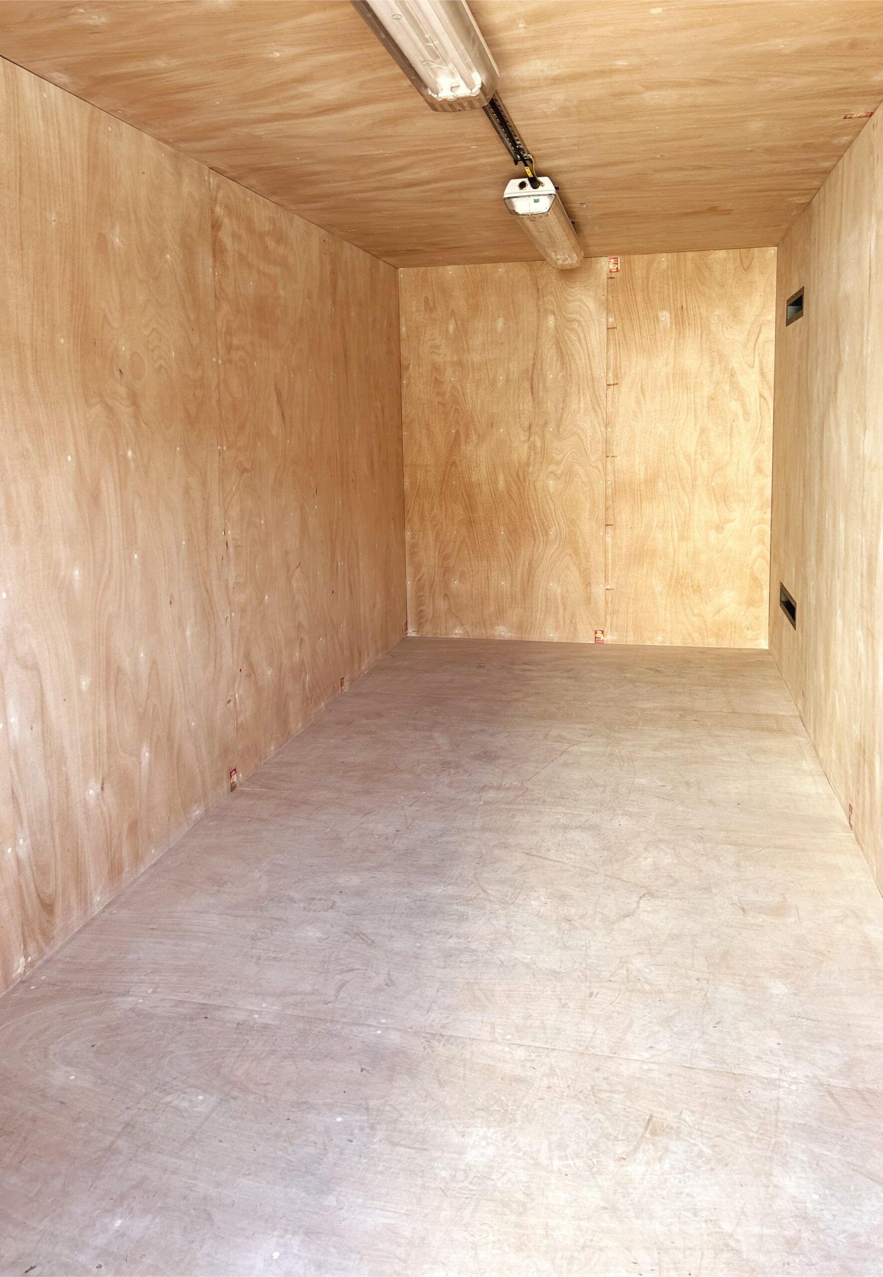 Empty storage unit with plywood walls and concrete floor, illuminated by fluorescent lights.