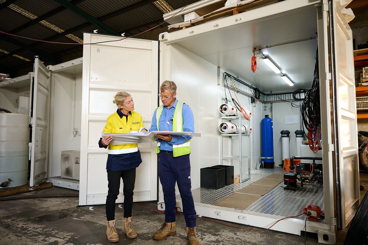 Two workers in high-visibility jackets standing beside an open industrial container with equipment inside