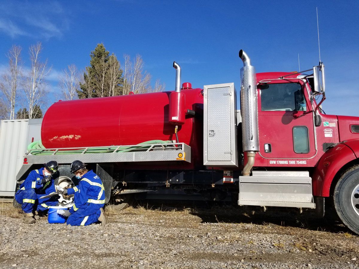 Two Centurion workers in blue gear crouch beside a red sewage truck on gravel under a clear blue sky