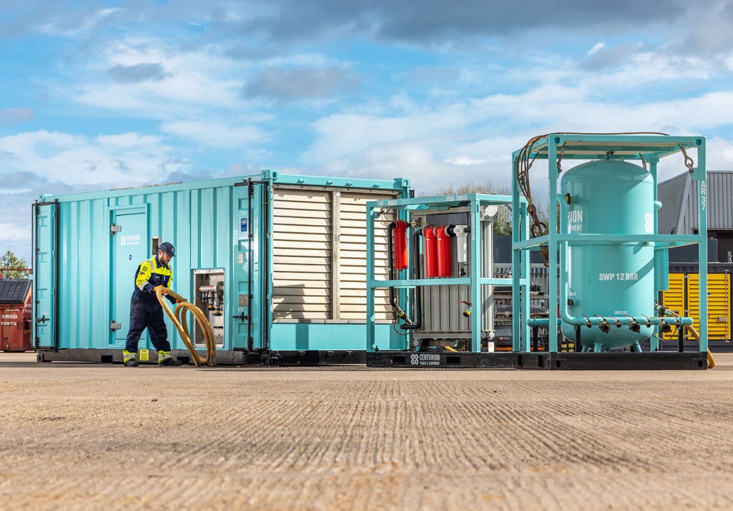 Man in work clothes near a turquoise container with machinery, outside on a sunny day.