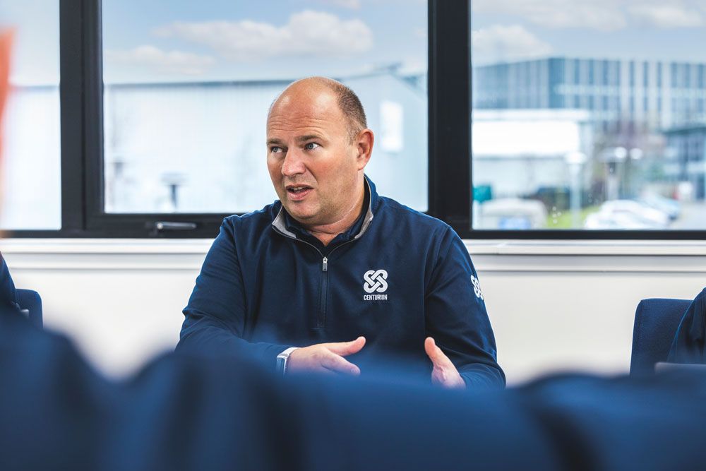 Man in blue jacket gesturing while speaking in a meeting, with windows and modern building in background.