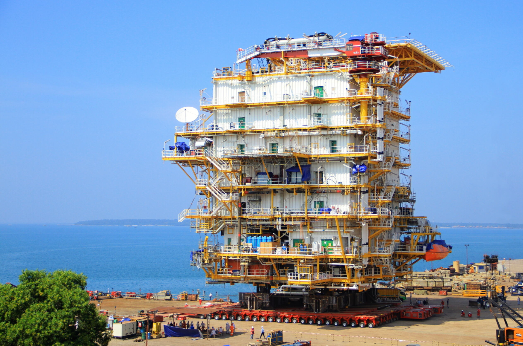 Large, multi-story industrial structure on wheeled transporters near a body of water, under a blue sky.
