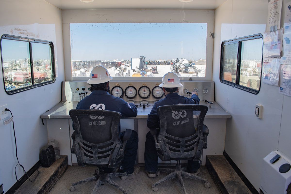 Two workers in hard hats monitor controls inside a white control room, overlooking an industrial site.