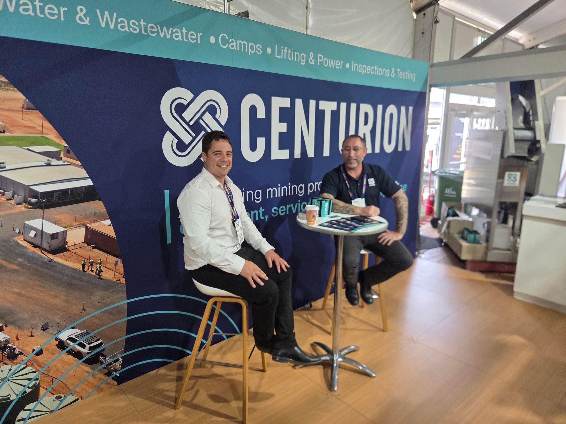 Two people seated at a Centurion booth at a trade show, with a large blue banner behind them.