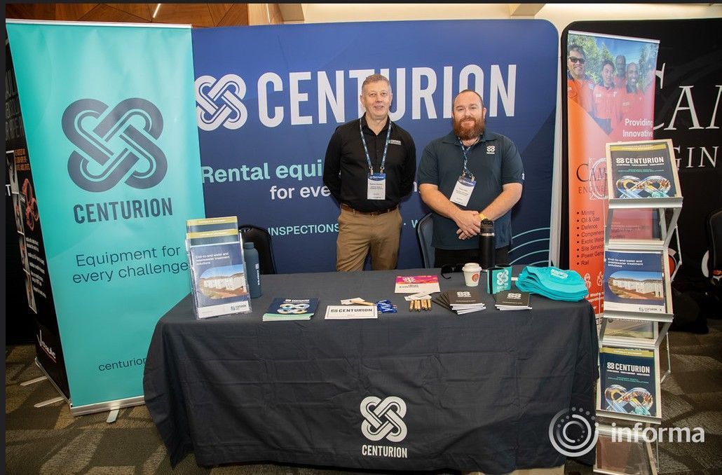 Two people standing at a Centurion booth at a trade show, with a large blue banner behind them and a green banner beside them.