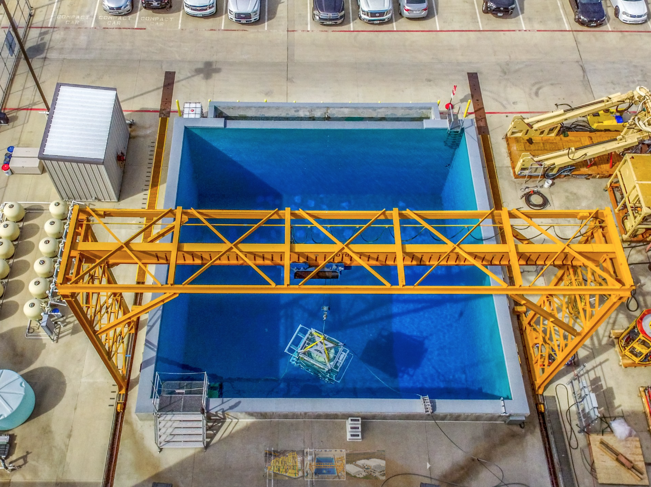 Overhead view: Large square blue pool with yellow gantry crane; equipment and vehicles nearby.